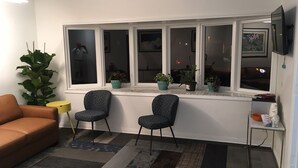 Ein Wohnzimmer mit einer Couch, zwei gemusterten Sesseln, einem kleinen Tisch und einem Fenster mit Blumentöpfen.