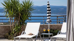 Une terrasse avec des fauteuils de jardin blancs, un parasol à rayures et une table sur laquelle sont posés deux verres de jus d’orange.