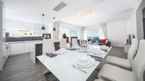 Un espace de repas moderne avec une table blanche dressée pour quatre personnes, une cuisine avec des meubles blancs et un salon doté d’un canapé gris et de coussins rouges.