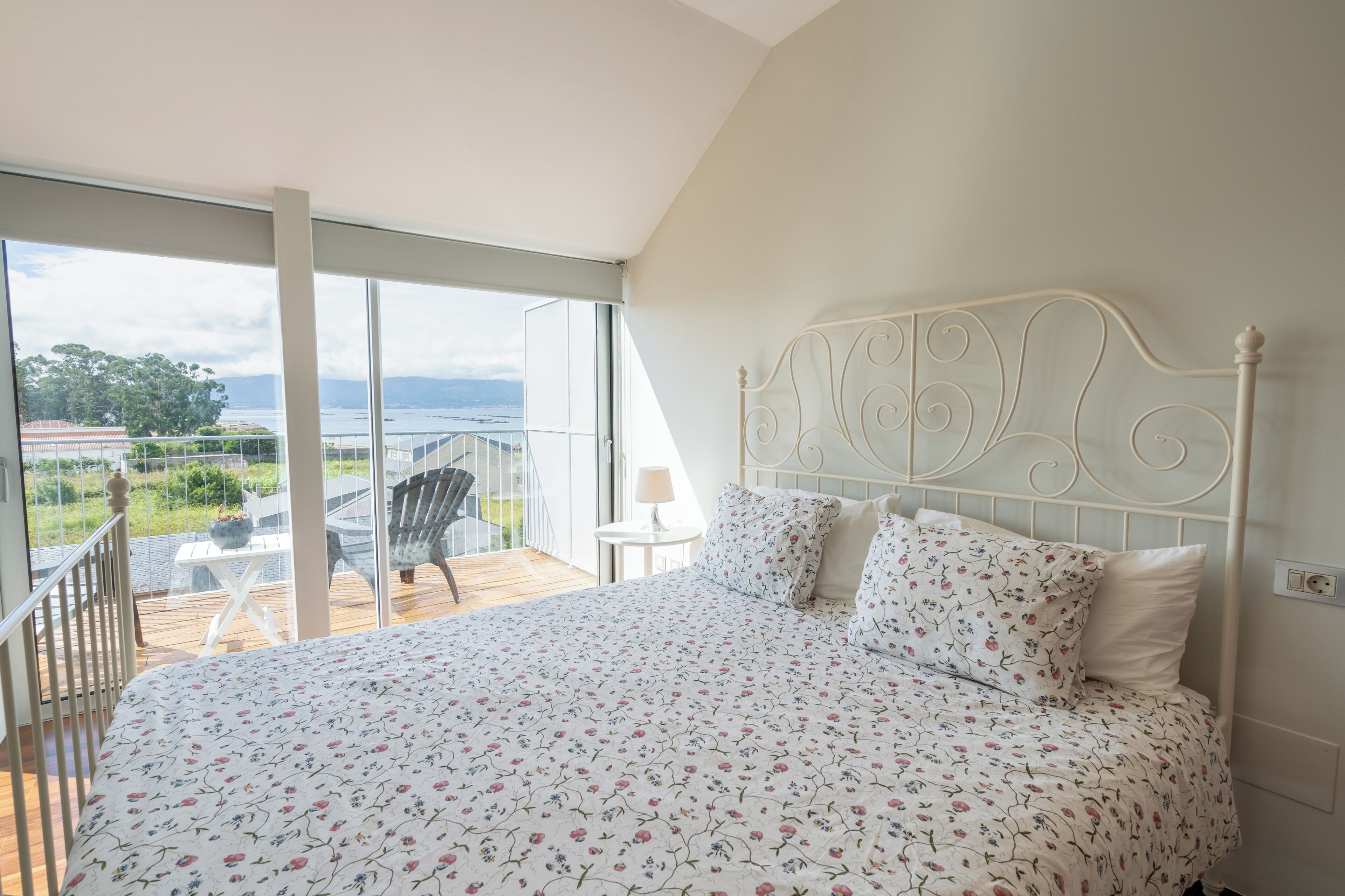 A bedroom with a large bed, a balcony with a chair, and a view of the sea.