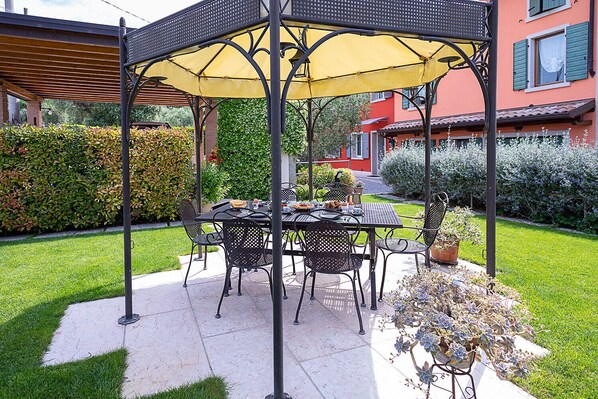 A covered outdoor dining area with a table and chairs, surrounded by a well-maintained garden and a building in the background. - Regarda – Petra holiday house in Bardolino, 3 bedrooms, garden and wifi (Bardolino)