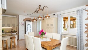 Un coin repas avec une table en bois, des chaises blanches et un lustre. Il y a également un coin cuisine avec des placards et une fenêtre.
