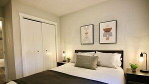 A bedroom with a bed, two bedside tables with lamps, a wardrobe, and two framed artworks on the wall. - Central, Modern and Cozy 1 Bedroom Retreat in the Heart of Tuxedo (Calgary)