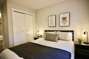 A bedroom with a bed, two bedside tables with lamps, a wardrobe, and two framed artworks on the wall. - Central, Modern and Cozy 1 Bedroom Retreat in the Heart of Tuxedo (Calgary)