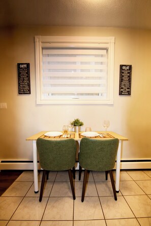 A dining area with a table set for two, two chairs, a window with blinds, and two wall signs listing coffee and gelato options. - Central, Modern and Cozy 1 Bedroom Retreat in the Heart of Tuxedo (Calgary)