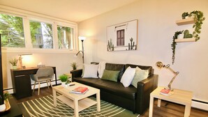 A living room with a black leather sofa, a wooden coffee table, a desk with a chair, and a lamp. - Central, Modern and Cozy 1 Bedroom Retreat in the Heart of Tuxedo (Calgary)