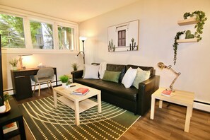 A living room with a black leather sofa, a wooden coffee table, a desk with a chair, and a lamp. - Central, Modern and Cozy 1 Bedroom Retreat in the Heart of Tuxedo (Calgary)
