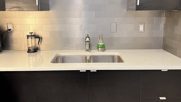 A modern kitchen with dark cabinets, a stainless steel sink, and a tiled backsplash. - Central, Modern and Cozy 1 Bedroom Retreat in the Heart of Tuxedo (Calgary)