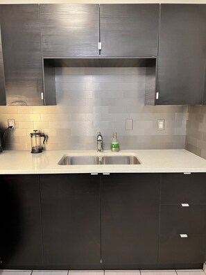 A modern kitchen with dark cabinets, a stainless steel sink, and a tiled backsplash. - Central, Modern and Cozy 1 Bedroom Retreat in the Heart of Tuxedo (Calgary)