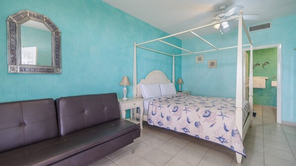 Uma cama de dossel com padrão de estrela-do-mar e conchas, um sofá marrom-escuro, um ventilador de teto branco e um espelho com moldura decorativa.