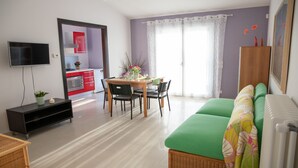 Ein modernes Wohnzimmer mit einem grünen Sofa, einer Essecke mit Tisch und Stühlen, einer Küche mit roten Schränken und einem an der Wand montierten Fernseher.