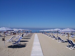 Una platja amb files de gandules i ombrel·les, cel blau clar i una passarel·la que condueix a l'aigua.