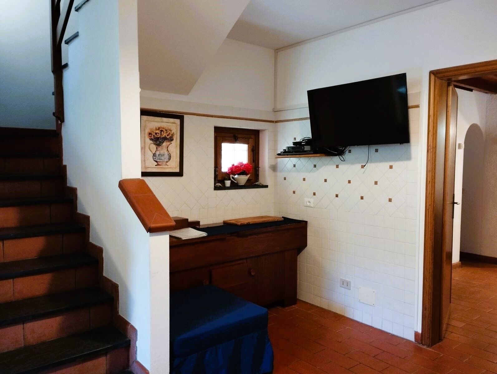 Una cocina con una escalera de madera, un televisor de pantalla plana montado en la pared y una ventana pequeña con un florero.