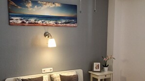 Une chambre à coucher avec un lit, un ventilateur de plafond, une lampe fixée au mur, une petite table sur laquelle se trouve un cadre photo et un tableau représentant une scène de plage.