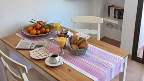 Une table préparée pour le petit-déjeuner, avec des chaises, un panier d’oranges, un verre de jus d’orange, un panier de pain et un téléviseur fixé au mur.