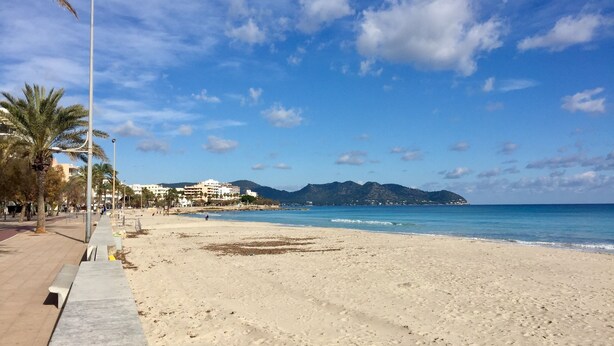 Une vaste plage de sable fin, sous un ciel bleu azur, avec des montagnes qui se dessinent à l’horizon.
