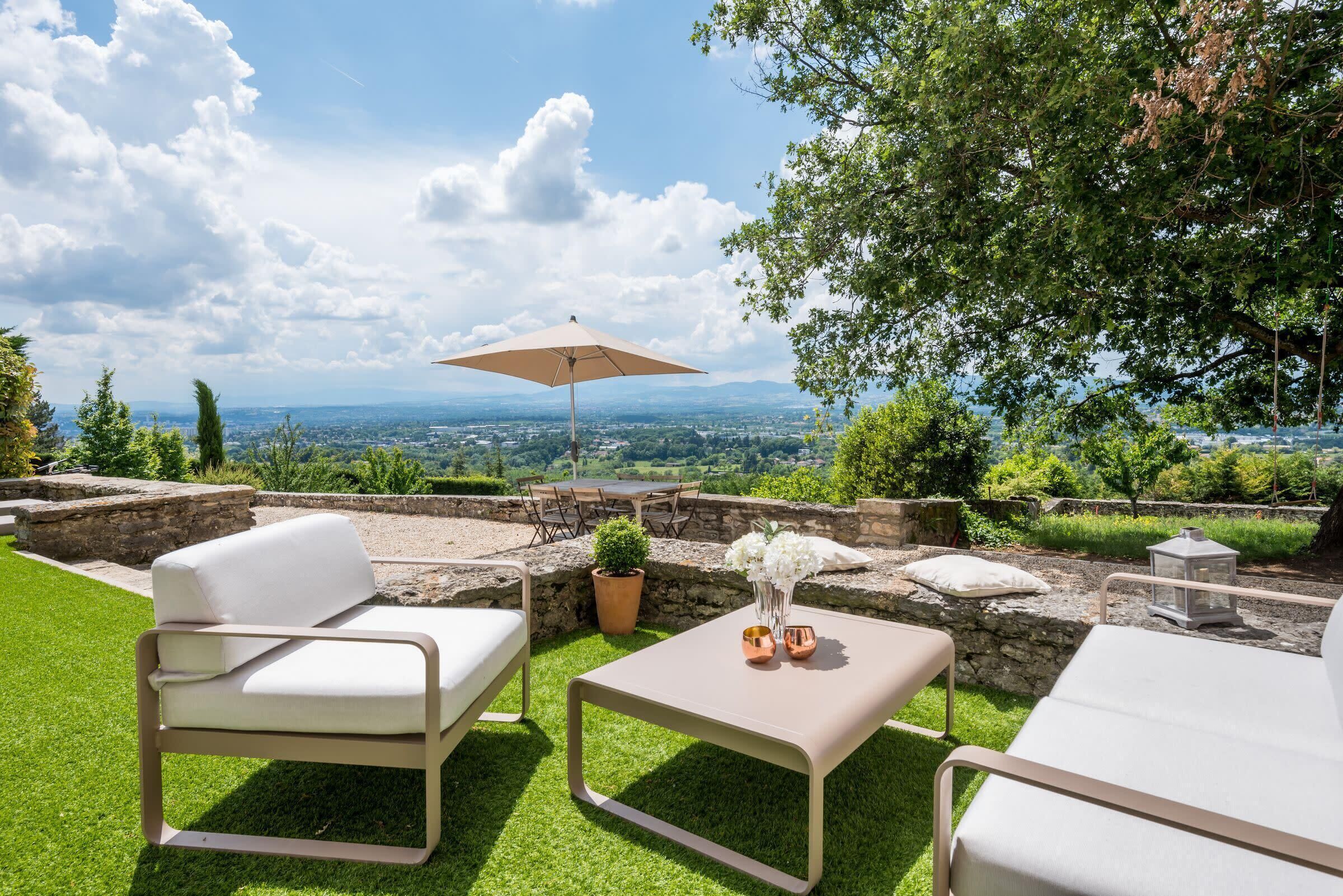 An outdoor patio with a white sofa, a table, and an umbrella. There is a view of a cityscape in the background.