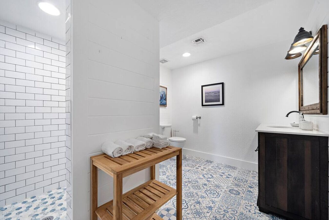 A bathroom with white tiled walls, a wooden vanity, a sink, a toilet, and a shower area.