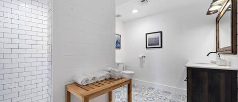 A bathroom with white tiled walls, a wooden vanity, a sink, a toilet, and a shower area.
