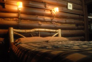A log cabin room with a wooden bed, two wall-mounted lamps, and a framed picture on the wall.