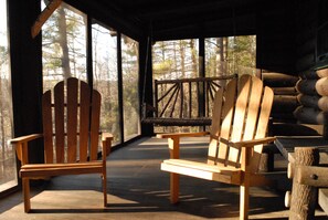 A wooden cabin with two Adirondack chairs facing a wooden deck and a forest view.