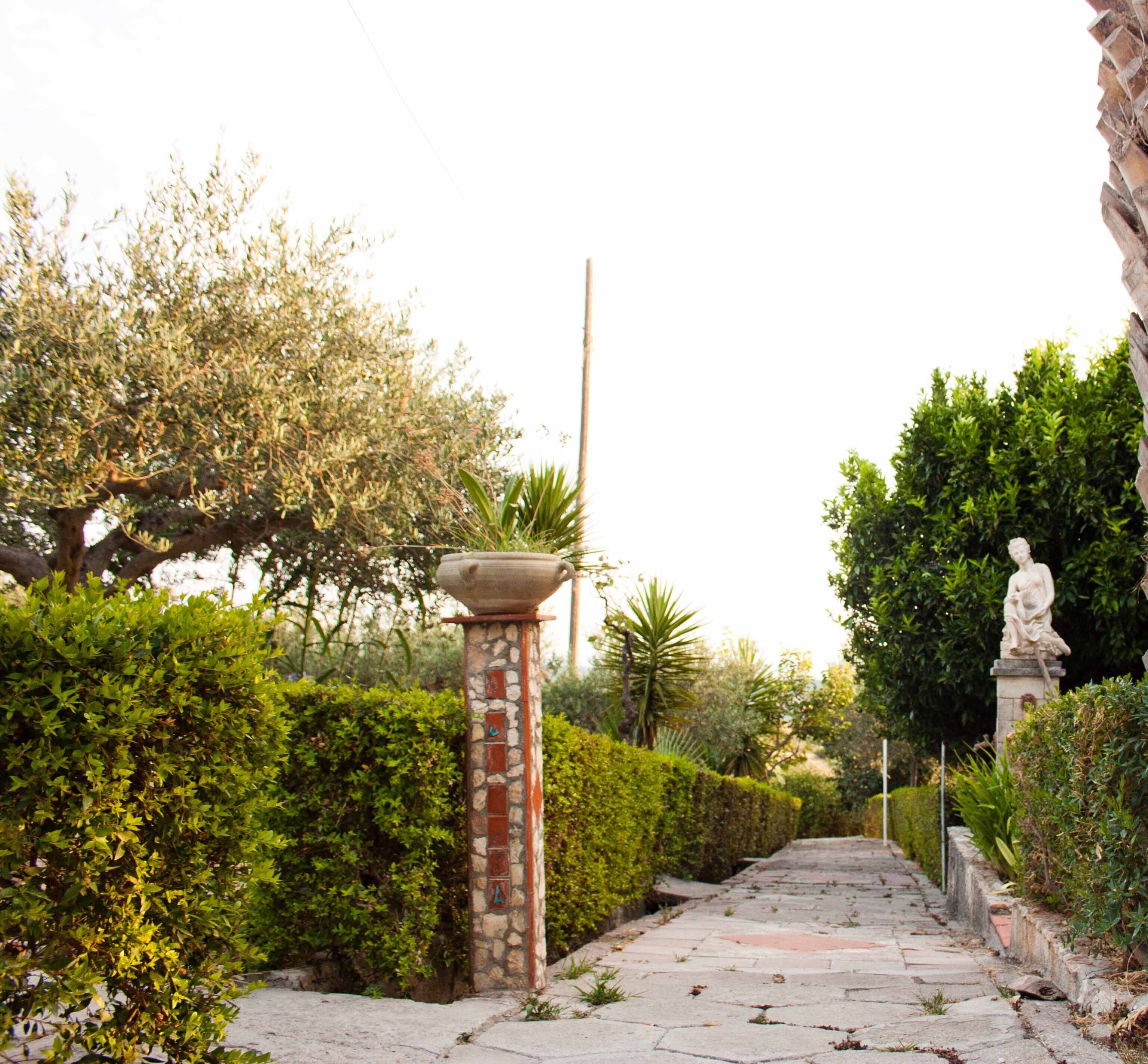 A stone pathway leading through a garden with a statue and a fountain.