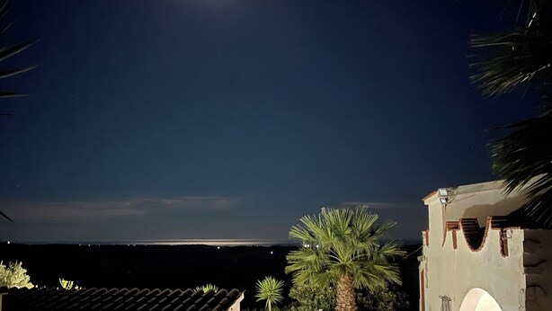 Une vue nocturne d’une cour avec un sol carrelé, un palmier et un bâtiment à la façade en pierre.