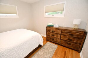 A bedroom with a bed, a dresser, two windows with blinds, and a rug.