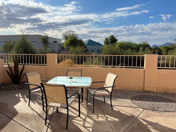 Un patio al aire libre con una mesa y sillas, una baranda de metal y vistas a edificios y árboles al fondo.