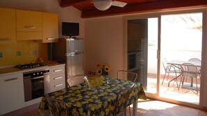 Une cuisine avec des meubles de couleur jaune, une cuisinière et un réfrigérateur. On y trouve une table à manger avec des chaises, ainsi qu’une fenêtre donnant sur un espace patio.