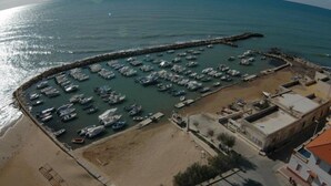 Vue aérienne d’une marina côtière où sont amarrés de nombreux bateaux, avec une plage de sable et quelques bâtiments à proximité.