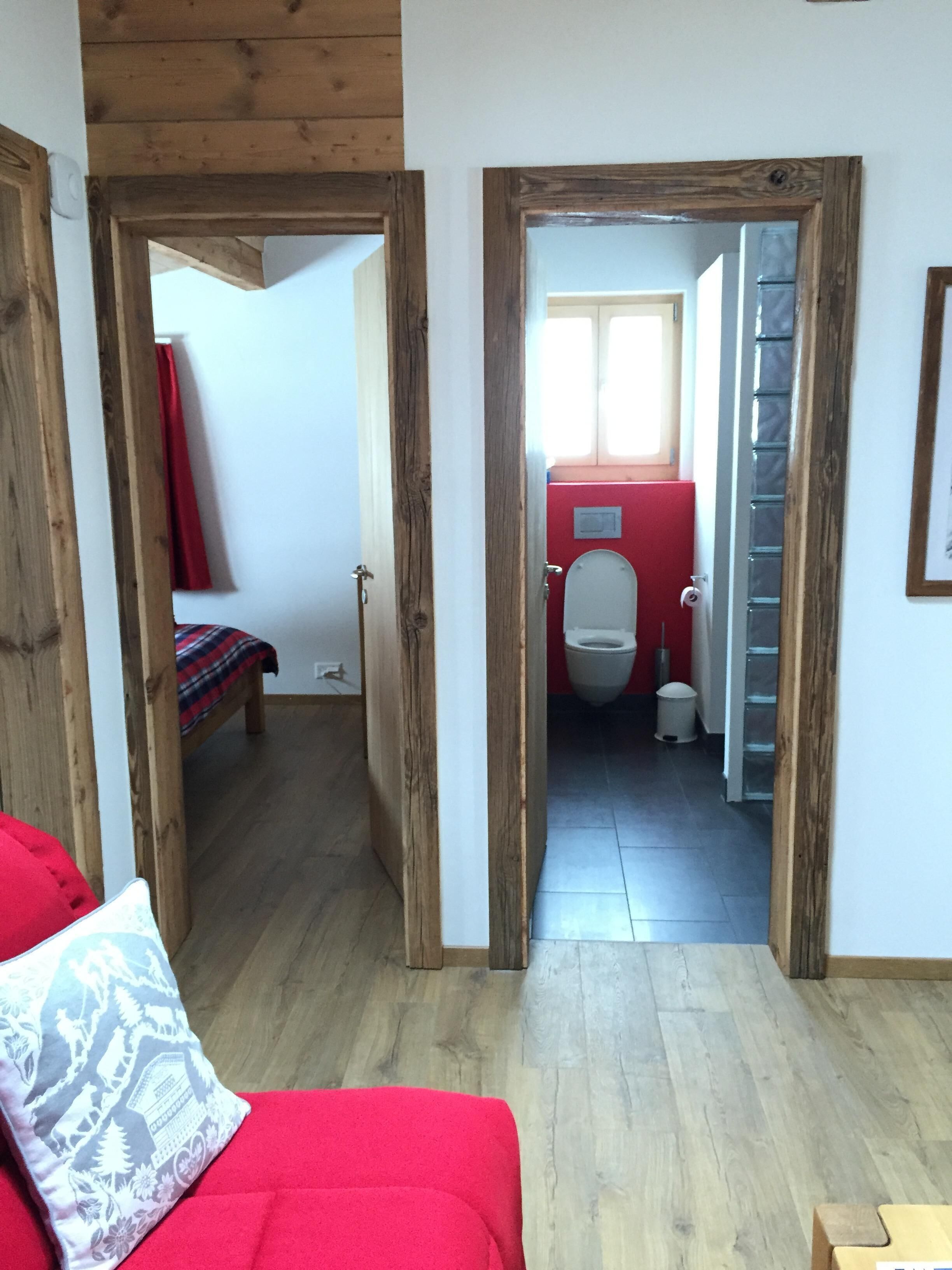 Une pièce avec un plancher en bois, un canapé rouge et une porte donnant sur une salle de bain équipée de toilettes et d’une poubelle.