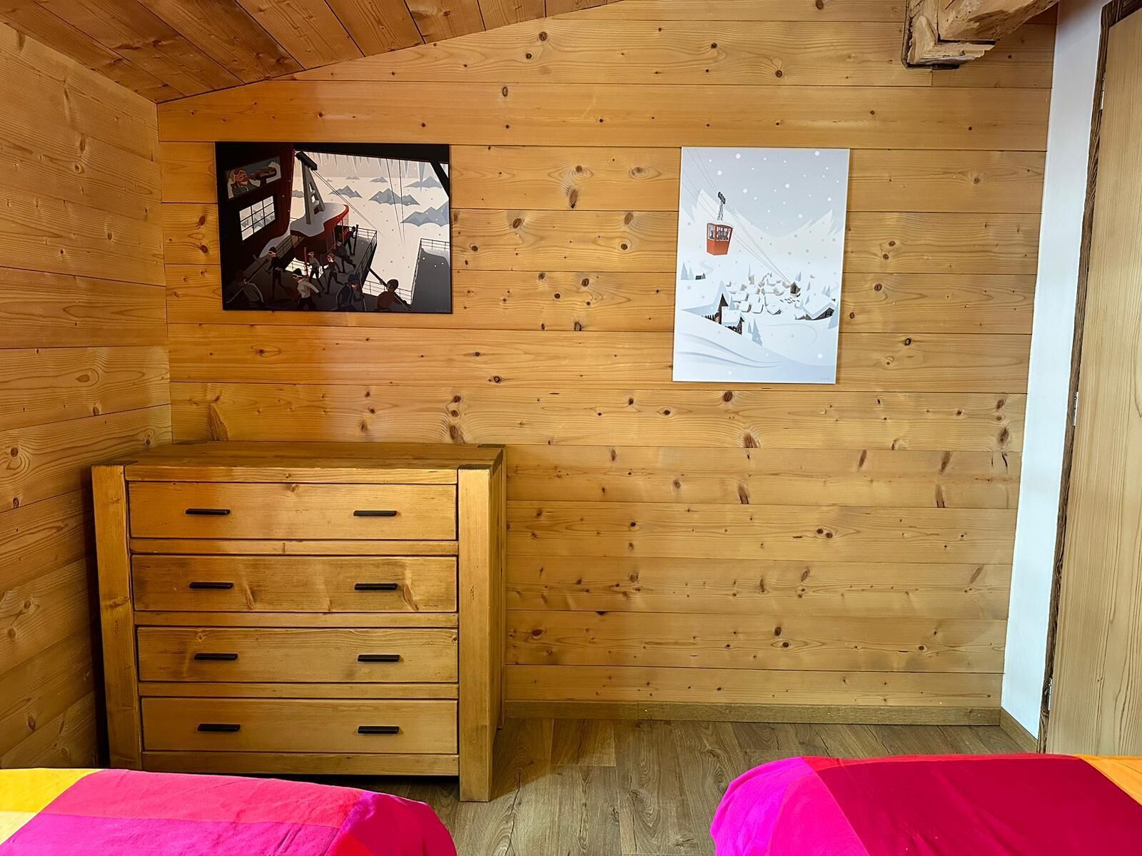 A wooden cabin room with a wooden dresser, two beds with pink bedding, and two wall-mounted pictures depicting snowy landscapes.