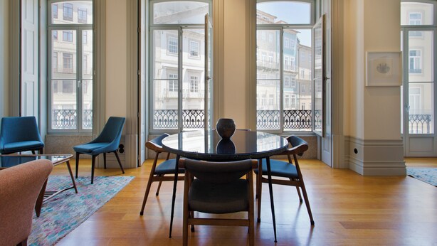 A dining area with a round table and chairs, large windows with a view of buildings, and a patterned rug on the floor.