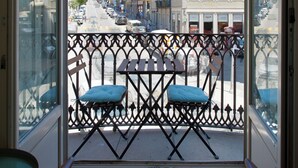A balcony with two blue cushioned chairs, a wrought iron railing, and a view of a city street with buildings and parked cars.