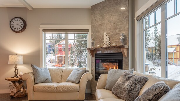 A cozy living room with a fireplace, two sofas, a lamp, and a view of a snowy landscape through large windows.