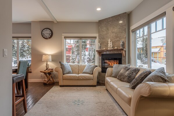 A cozy living room with two beige sofas, a fireplace, and large windows with a snowy view.