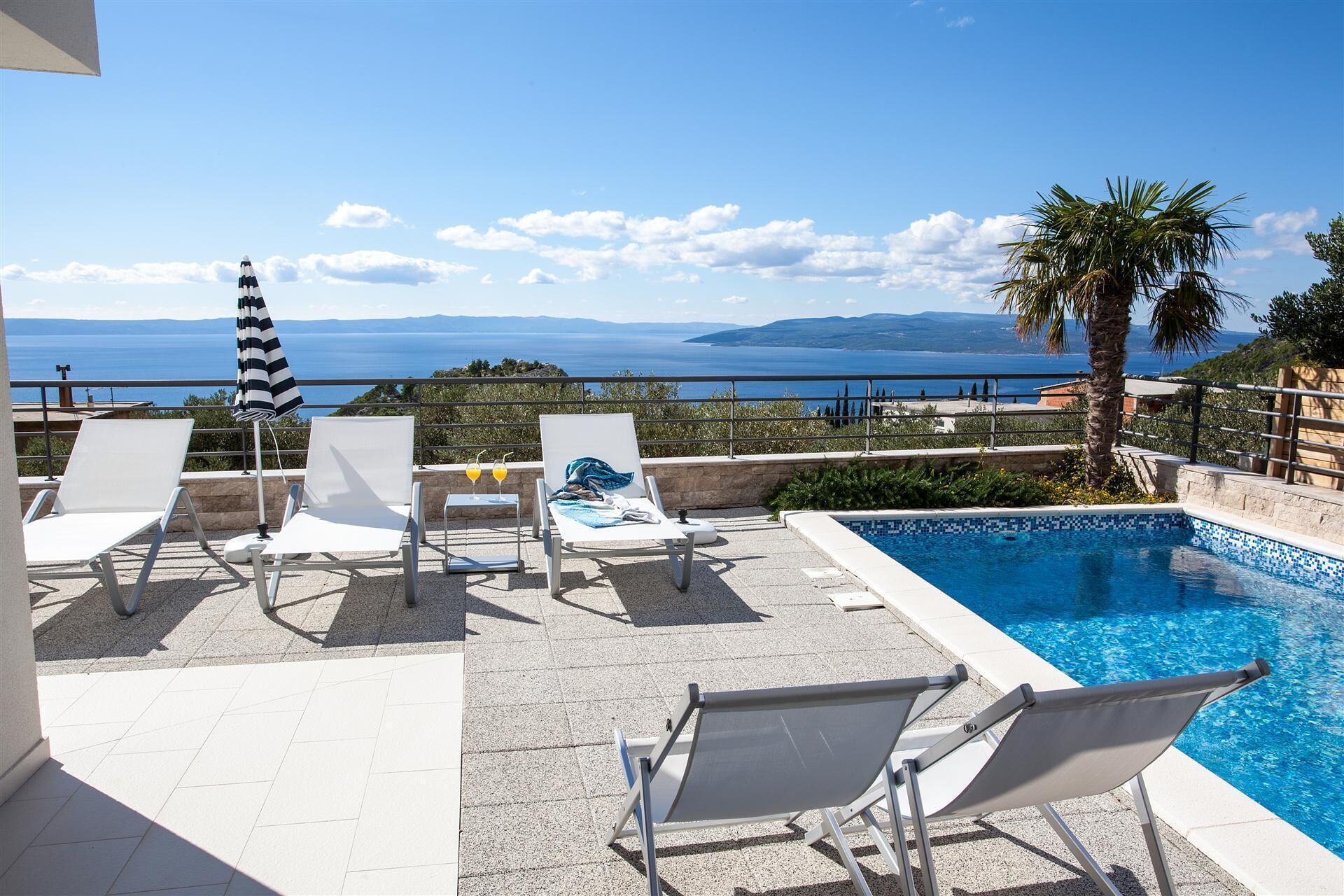 A poolside area with lounge chairs, a striped umbrella, and a view of the sea.