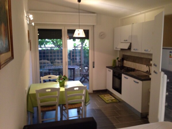 Cocina compacta con armarios blancos, estufa y refrigerador. Hay una mesa de comedor con sillas y una lámpara colgante.