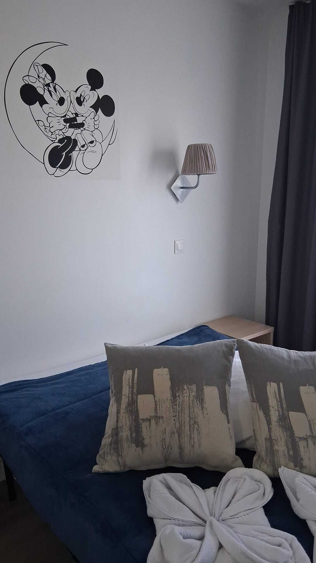 Une chambre à coucher avec un lit, des oreillers, une lampe fixée au mur et un autocollant mural représentant Mickey Mouse.