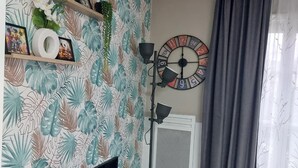 Un salon avec un téléviseur, une horloge, une plante et une fenêtre avec des rideaux.