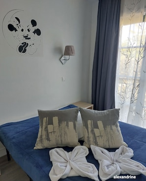 Une chambre à coucher avec un lit, des oreillers, une décoration murale représentant Mickey Mouse, une lampe et une fenêtre avec des rideaux.