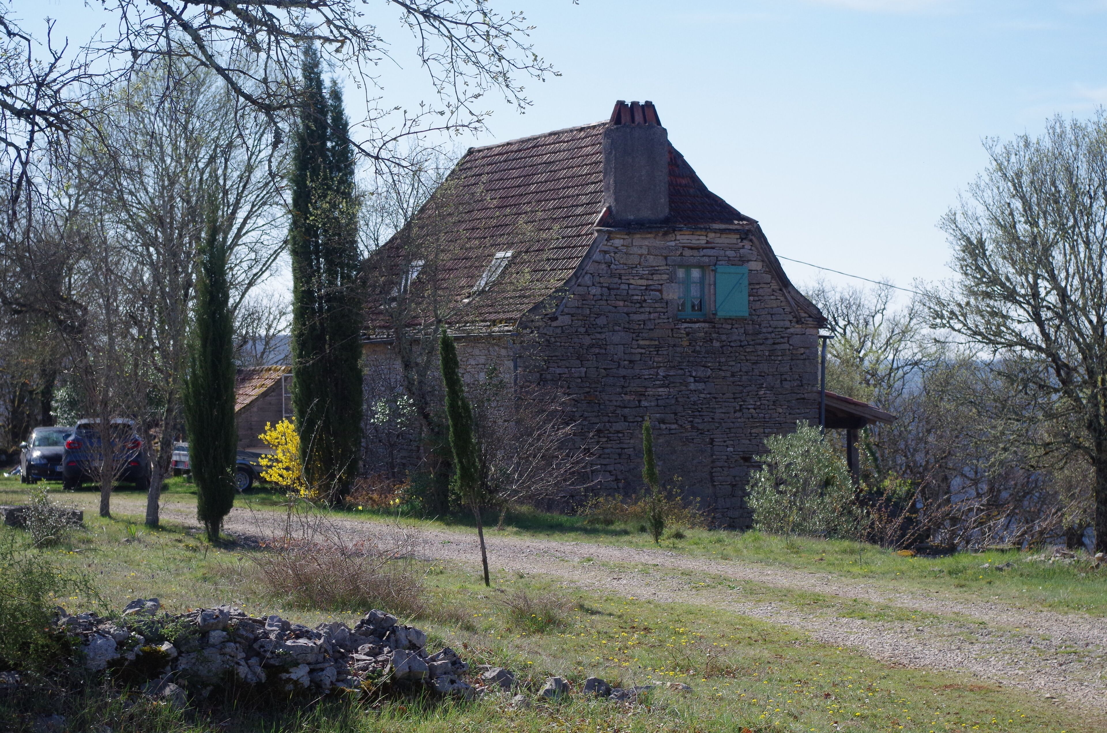 Une maison en pierre avec un toit en pente et une cheminée, entourée d’arbres et bordée d’un chemin de gravier.