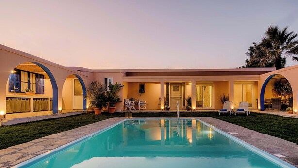 Una piscina con zona de patio, un área de estar techada y un edificio con grandes ventanales y puertas.