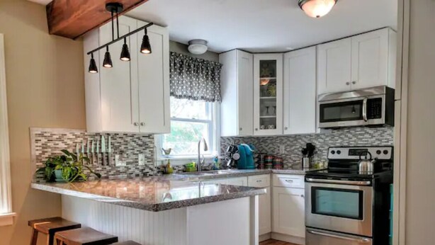 A kitchen with white cabinets, a granite countertop, a stainless steel oven, and a microwave. There are wooden stools and a window with patterned curtains. - ☆Oakley's Oasis, a Dharma Dwellings Designed Home☆ (Rochester)