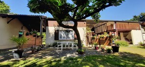 Une maison dotée d’un patio couvert, d’un petit jardin avec une table et des chaises, et de diverses plantes en pot.