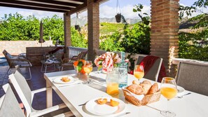 Un espace de restauration en plein air avec une table dressée pour le petit-déjeuner, comprenant des fruits, du pain et des boissons.