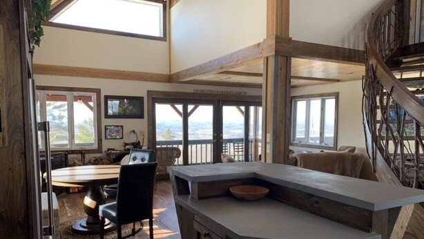 A spacious living area with a large wooden dining table, a built-in kitchen island, and a view of the outdoors through large windows. - A soul soothing escape. Artistically built and lovingly maintained. (New Sharon)