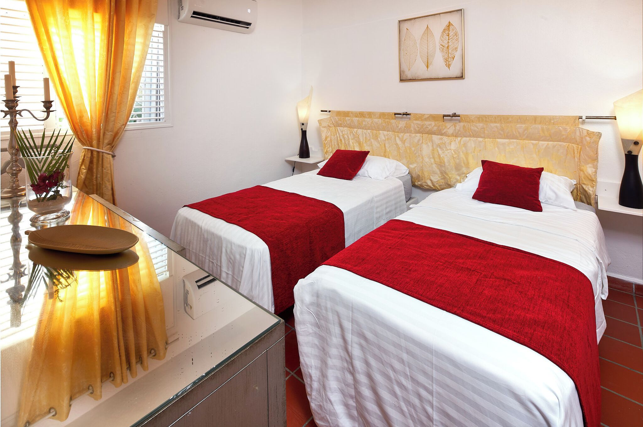 A hotel room with two beds, each with a red and white bedspread, a bedside table with a lamp, and a window with curtains.