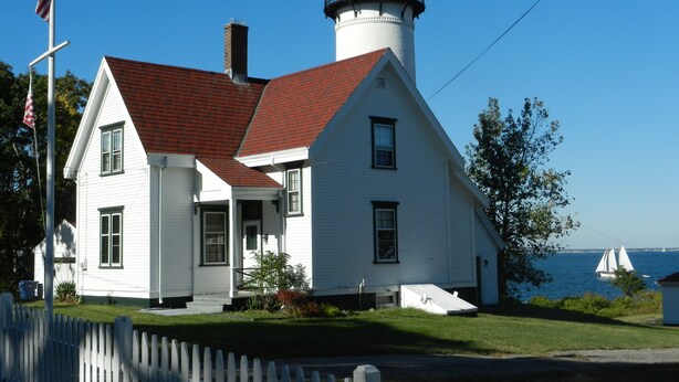 Um edifício branco com telhado vermelho e uma torre de farol, situado perto de um corpo de água.